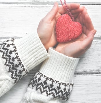 Red heart in the hands of a young woman in a white wool sweater with an ornament on a white wooden rustic background.